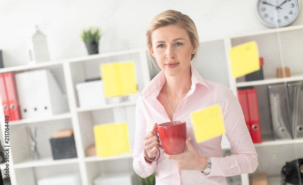 A young blonde girl stands in the office next to a transparent board with stickers and holds a red cup in her hands. A young blonde girl stands in the office next to a transparent board with stickers and holds a red cup in her hands.