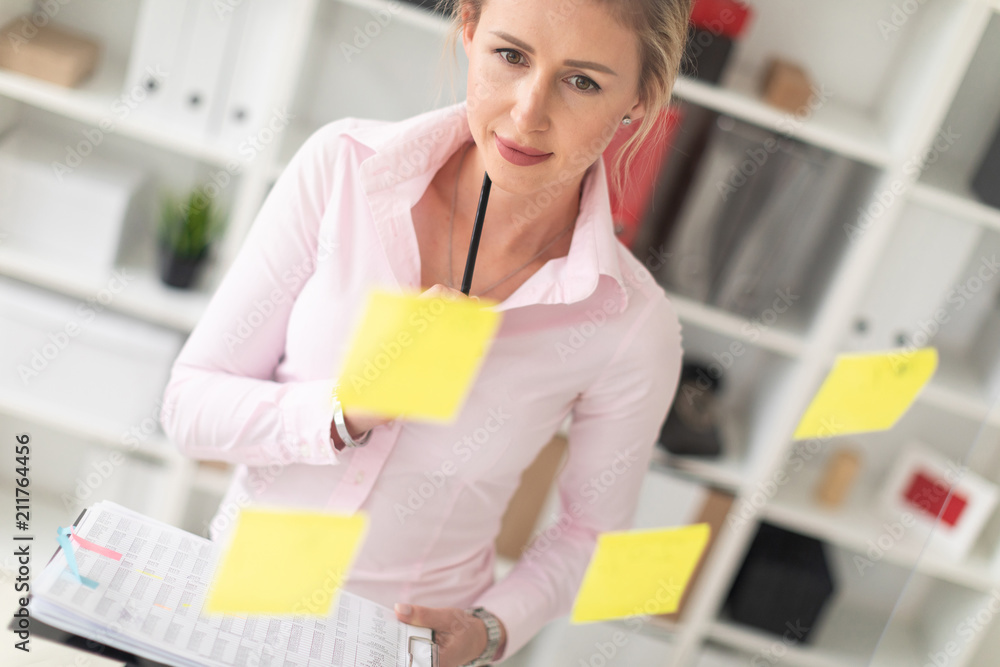 A young blonde girl stands in the office next to a transparent board with stickers and holds documents and a pencil in her hands. A young blonde girl stands in the office next to a transparent board with stickers and holds documents and a pencil in her hands.