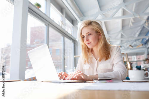 Charming businesswoman writes an article in cafe sitting at table with laptop and documents and using high-speed internet