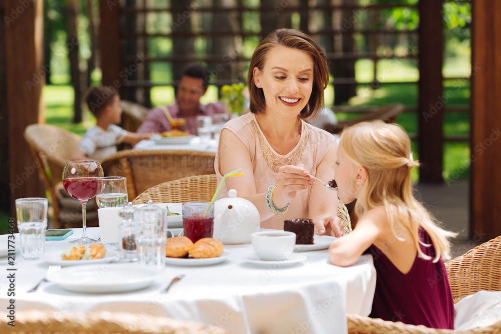 Birthday cake. Little blonde-haired girl feeling extremely happy while eating her birthday cake sitting near mother Birthday cake. Little blonde-haired girl feeling extremely happy while eating her birthday cake sitting near mother