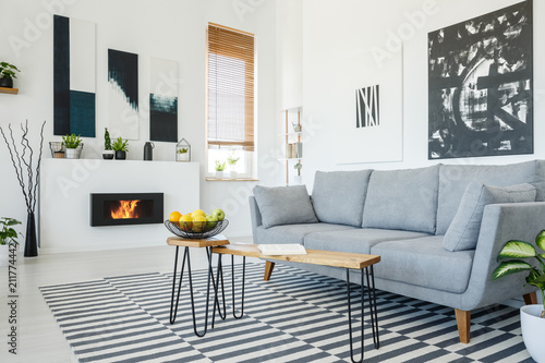 Real photo of a grey sofa standing in front of a wooden table in living room interior with posters on the walls, striped rug and bio fireplace