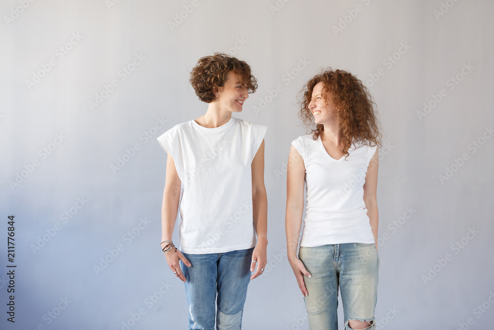 Studio shot of two European sisters with curly hair, wearing white t-shirts and jeans, smiling happily, looking at each other, posing at gray wall. People and friendship concept Studio shot of two European sisters with curly hair, wearing white t-shirts and jeans, smiling happily, looking at each other, posing at gray wall. People and friendship concept