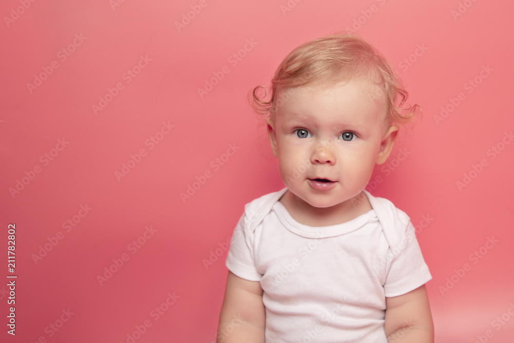 Naklejka premium Portrait of cute adorable Caucasian baby girl with blue eyes and curly hair on pink background looking in camera.