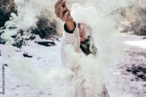 .Young and relaxed woman enjoying a sunny winter day in the mountains. Surrounded by snow and trunks holding a tube of green smoke, funny photo. Lifesytle