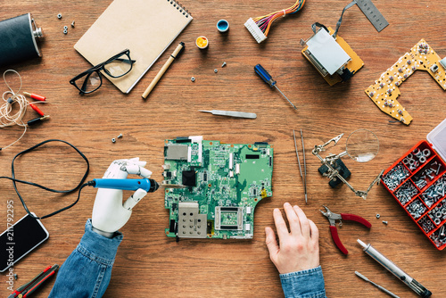 cropped image of electronic engineer with robotic hand fixing motherboard by soldering iron at table surrounded by tools