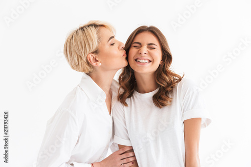Portrait closeup of two caucasian lovely women, adult mother kissing on cheek her teenage daughter, standing isolated over white background