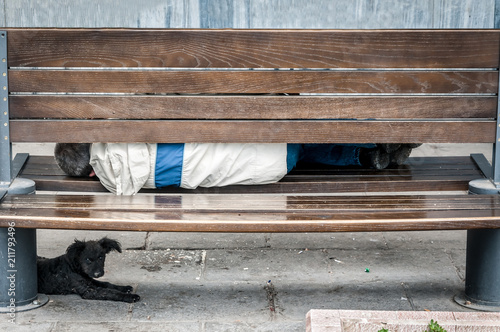 Poor homeless man with his dog sleeping on the urban street in the city on the wooden bench. Homeless people concept.