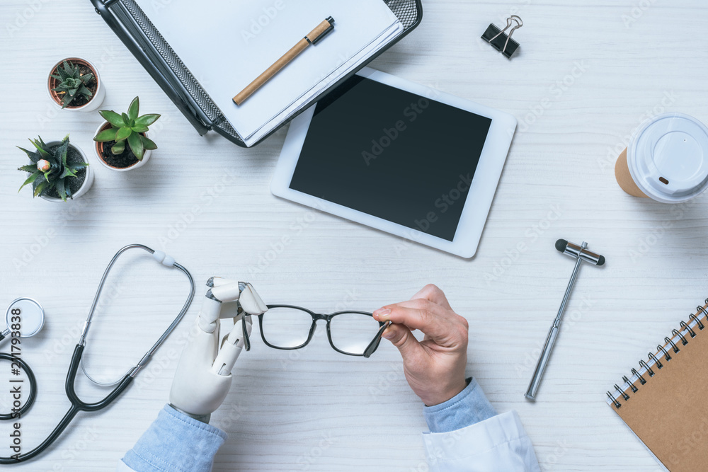 cropped image of male doctor with prosthetic arm holding eyeglasses at table with blank digital tablet cropped image of male doctor with prosthetic arm holding eyeglasses at table with blank digital tablet
