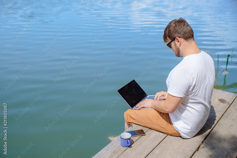 man working on laptop while sitting on wooden dock. legs in river water