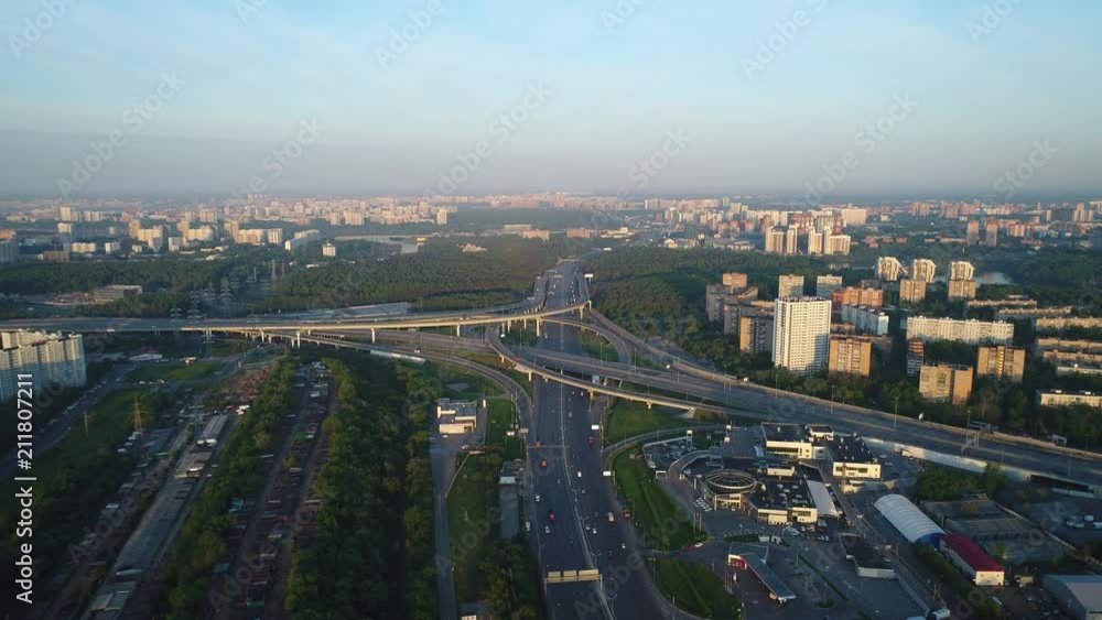 Aerial view. Flight over a huge interchange of the Moscow ring road in the early morning at dawn from high altitude. Cars are moving on a multi-level road junction. Big city in background. Aerial view. Flight over a huge interchange of the Moscow ring road in the early morning at dawn from high altitude. Cars are moving on a multi-level road junction. Big city in background.