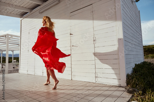 Outdoor portrait of dancing blonde woman in wavy red dress with her shadow on white wooden building near her. Mountains on background photo