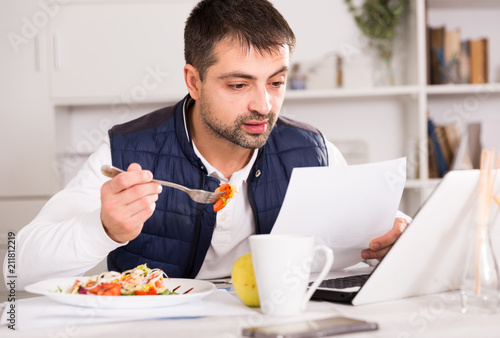 Young man eating vegetable salad and working at laptop