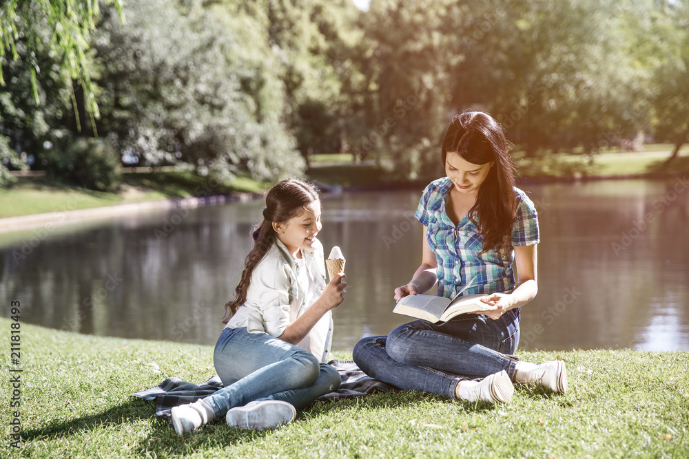 Smart woman is sitting at the edge of small lake with her daughter and reading a book. Her kid is sitting besides her and eating ice cream. Girl is looking at what mom is doing right now. Smart woman is sitting at the edge of small lake with her daughter and reading a book. Her kid is sitting besides her and eating ice cream. Girl is looking at what mom is doing right now.