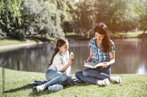 Smart woman is sitting at the edge of small lake with her daughter and reading a book. Her kid is sitting besides her and eating ice cream. Girl is looking at what mom is doing right now.