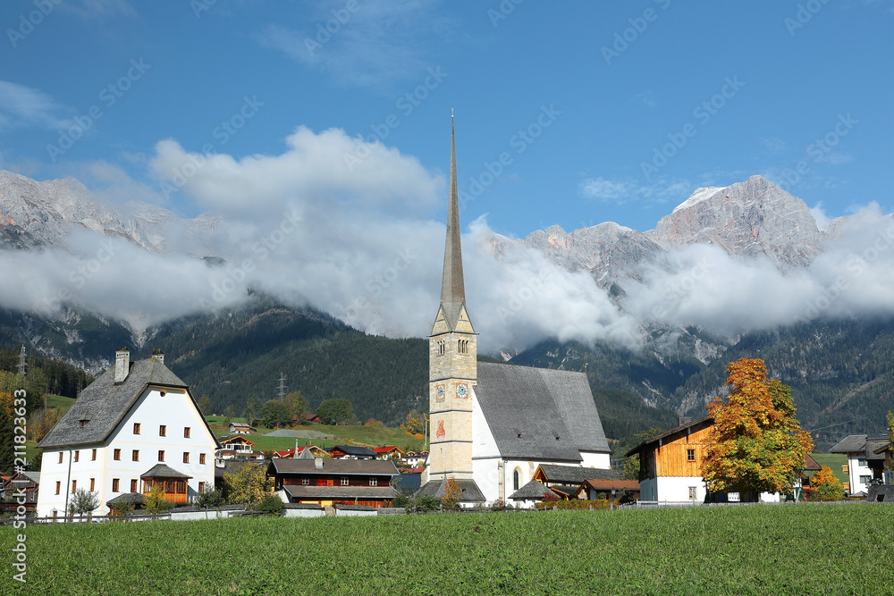 A peaceful village at the foothills of Steinernes Meer Mountains in Maria Alm, Austria, with a majestic church tower (Wallfahrtskirche) standing tall in green fields & foggy mountains in background