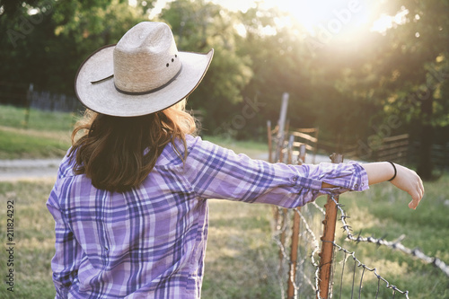 Western rural farm scene with cowgirl in cowboy hat and plaid shirt overlooking pasture during sunset.  Strong woman in country lifestyle.