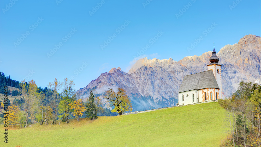 A church standing high on a grassy hilltop with a rocky mountain range in background~ Autumn scenery of St. Nikolaus Parish Church of Dienten Village at the foothill of Hochkoenig Mountains in Austria