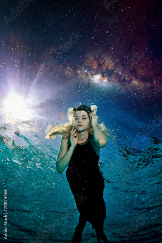 Young beautiful girl with white horns on her head swims in a black dress at night underwater in the pool against the background of space, the night sky and stars. She's looking at the camera. © alexbard