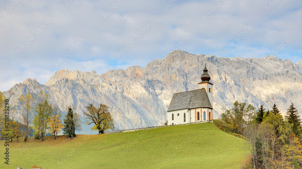 A church standing high on a grassy hilltop with a rocky mountain range in background~ Autumn scenery of St. Nikolaus Parish Church of Dienten Village at the foothill of Hochkoenig Mountains in Austria A church standing high on a grassy hilltop with a rocky mountain range in background~ Autumn scenery of St. Nikolaus Parish Church of Dienten Village at the foothill of Hochkoenig Mountains in Austria