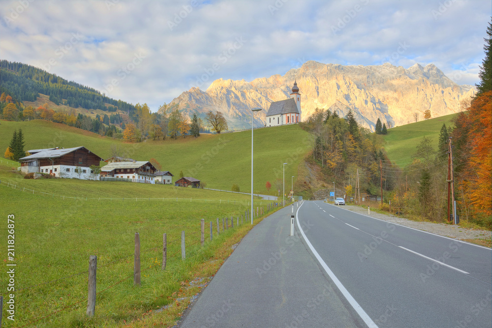 Scenic view of a country road passing by a farm land with a church on hilltop and rocky Hochkonig Mountain lighted up by alpenglow in the background in Dienten, Austria ~ Beautiful countryside scenery