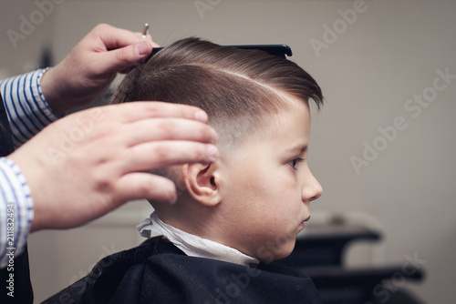 Caucasian boy getting hairstyle by hairdresser in barbershop. Side view.