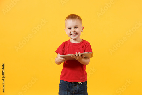 Little cute kid baby boy 3-4 years old in red t-shirt holding in hand tablet pc computer isolated on yellow background. Kids childhood lifestyle concept. Problem of children and gadgets. Copy space.