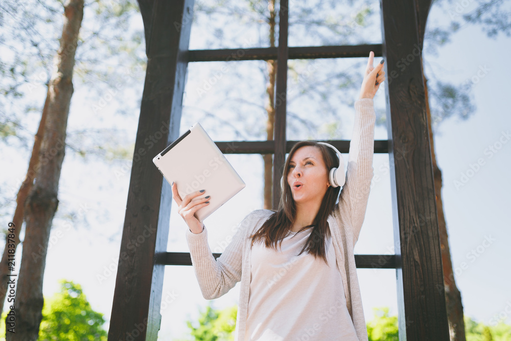 Naklejka premium Portrait of beautiful fun woman wearing light casual clothes. Pretty girl holding tablet pc computer, listening music in headphones in city park in street outdoors on spring nature. Lifestyle concept.