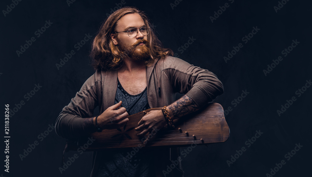 Redhead hipster male with long luxuriant hair and full beard dressed in casual clothes playing on a Russian traditional musical instrument - gusli. Isolated on a dark background.