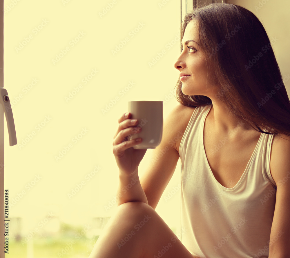 Beautiful smiling positive woman sitting near the window and looking up holding the cup of coffee in hand. Closeup toned color portrait Beautiful smiling positive woman sitting near the window and looking up holding the cup of coffee in hand. Closeup toned color portrait