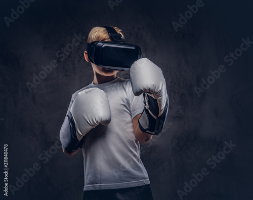 Fototapeta Naklejka Na Ścianę i Meble -  Schoolboy boxer with blonde hair dressed in a white t-shirt wearing visual reality glasses and boxing gloves, workout in a studio. Isolated on dark textured background.