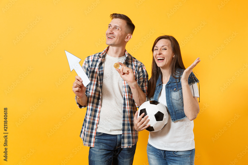 Fototapeta premium Couple, woman man, football fans holding bitcoin, metal golden coin, up arrow, soccer ball, cheer up support team isolated on yellow background. Sport bet, excitement ardor family lifestyle concept.