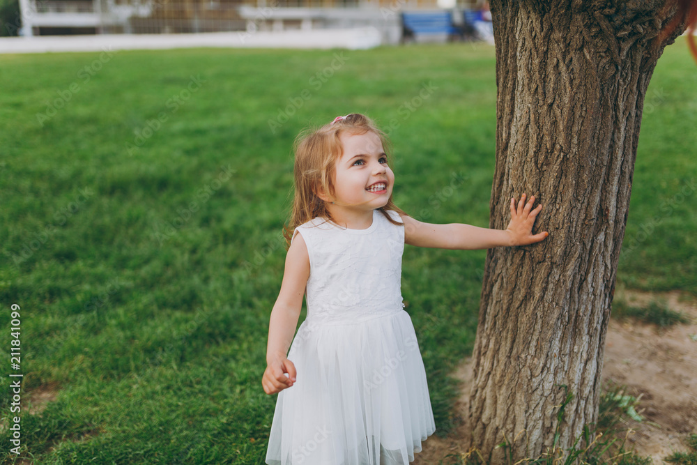 Naklejka premium Smiling little cute child baby girl in light dress stand and lean on tree on green grass in city park. Mother, little kid daughter. Mother's Day, love family, parenthood, childhood concept.
