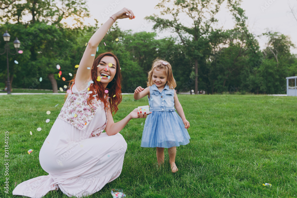Naklejka premium Woman in light dress and little cute child baby girl play with colorful paper confetti, have fun in green park. Mother, little kid daughter. Mother's Day, love family, parenthood, childhood concept.