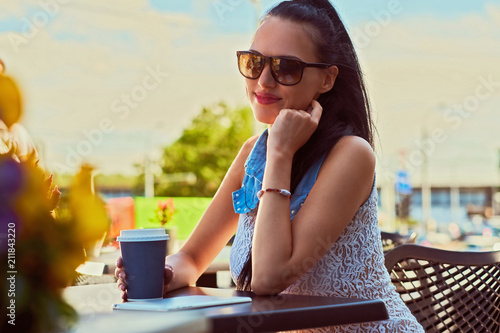 Close-up portrait of a beautiful brunette woman girl wearing trendy clothes is enjoying summer day while sitting on a terrace in an outdoor cafe.