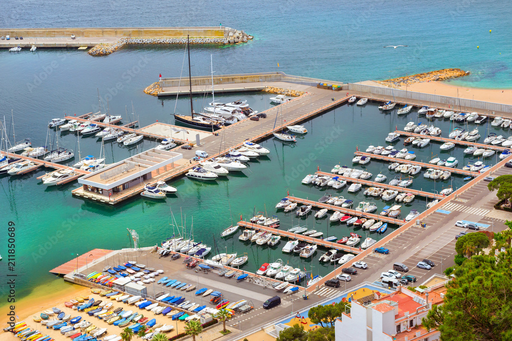 Sea port with boat berths and concrete promenade. Private yachts and fishing boats are moored at the pier. Coast of Spanish beach resort Blanes in summertime. Costa Brava, Catalonia, Spain