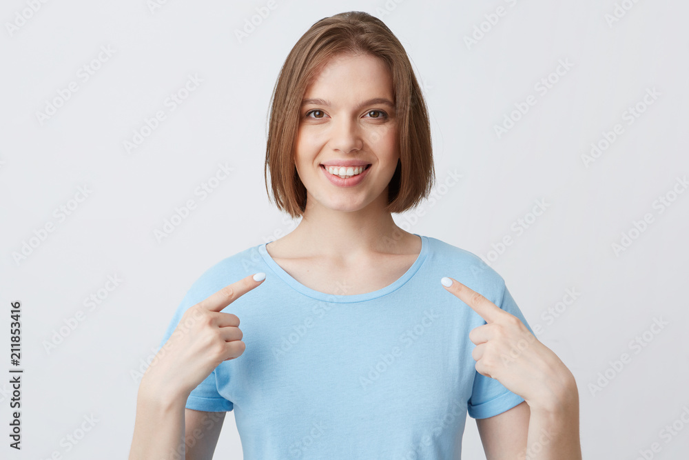 Smiling attractive young woman in blue t shirt feels confident and points at herself with fingers on both hands isolated over white background