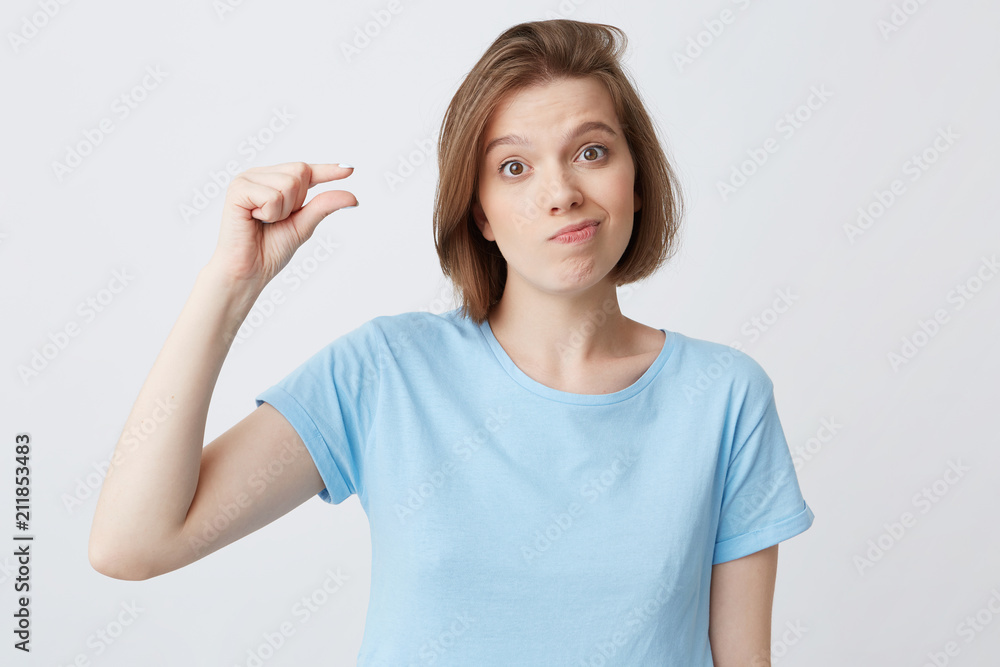 Unhappy disappointed young woman in blue t shirt frowning her face and showing tiny size by fingers isolated over white background Looks confused Unhappy disappointed young woman in blue t shirt frowning her face and showing tiny size by fingers isolated over white background Looks confused