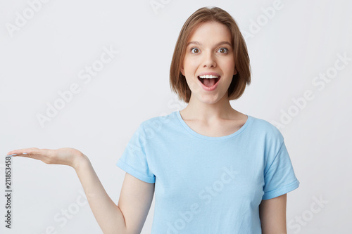 Wallpaper Mural Portrait of amazed excited young woman in blue t shirt standing with opened mouth and holding copyspace on palm isolated over white background Torontodigital.ca