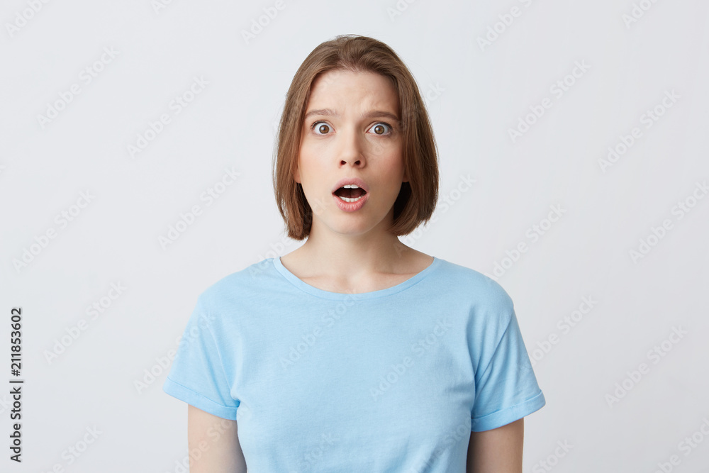 Portrait of amazed beautiful young woman in blue t shirt standing with opened mouth and feels shocked isolated over white background Portrait of amazed beautiful young woman in blue t shirt standing with opened mouth and feels shocked isolated over white background