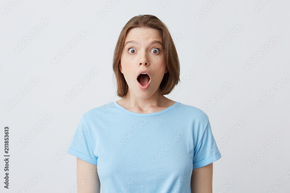 Closeup of shocked stunned young woman in blue t shirt with opened mouth feels amazed and looks directly in camera isolated over white background Closeup of shocked stunned young woman in blue t shirt with opened mouth feels amazed and looks directly in camera isolated over white background