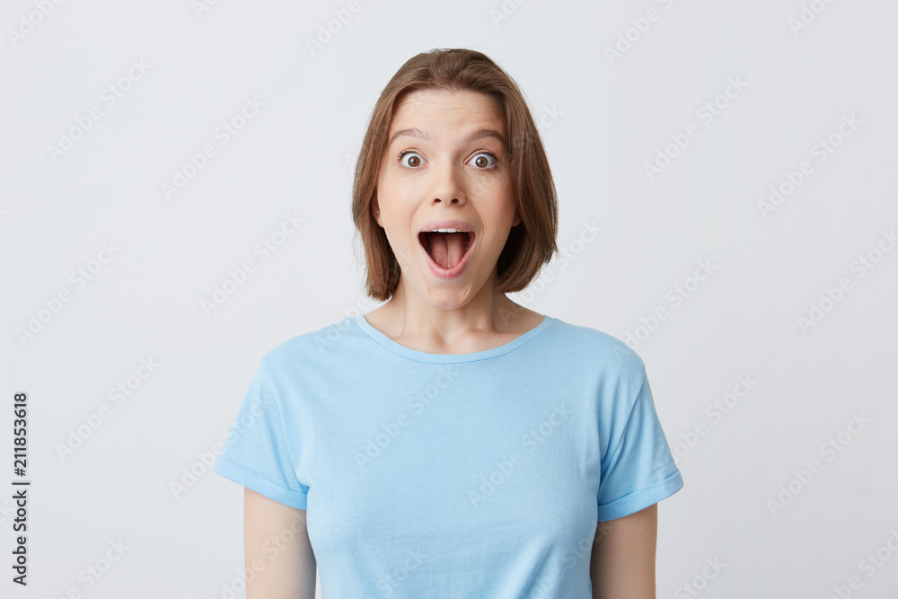 Portrait of surprised happy young woman with opened mouth in blue t shirt feels excited and shouting isolated over white background Portrait of surprised happy young woman with opened mouth in blue t shirt feels excited and shouting isolated over white background