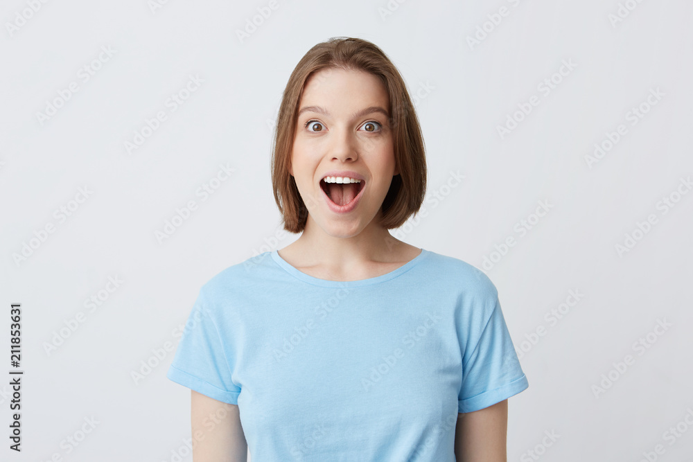 Closeup of amazed excited young woman with opened mouth in blue t shirt looks happy and shouting isolated over white background Feels excited Closeup of amazed excited young woman with opened mouth in blue t shirt looks happy and shouting isolated over white background Feels excited