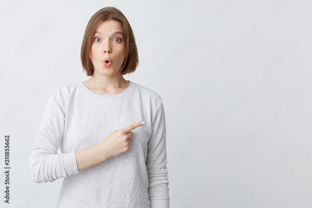 Amazed cute young woman in longsleeve making funny surprised face and pointing to the side with finger isolated over white background Amazed cute young woman in longsleeve making funny surprised face and pointing to the side with finger isolated over white background