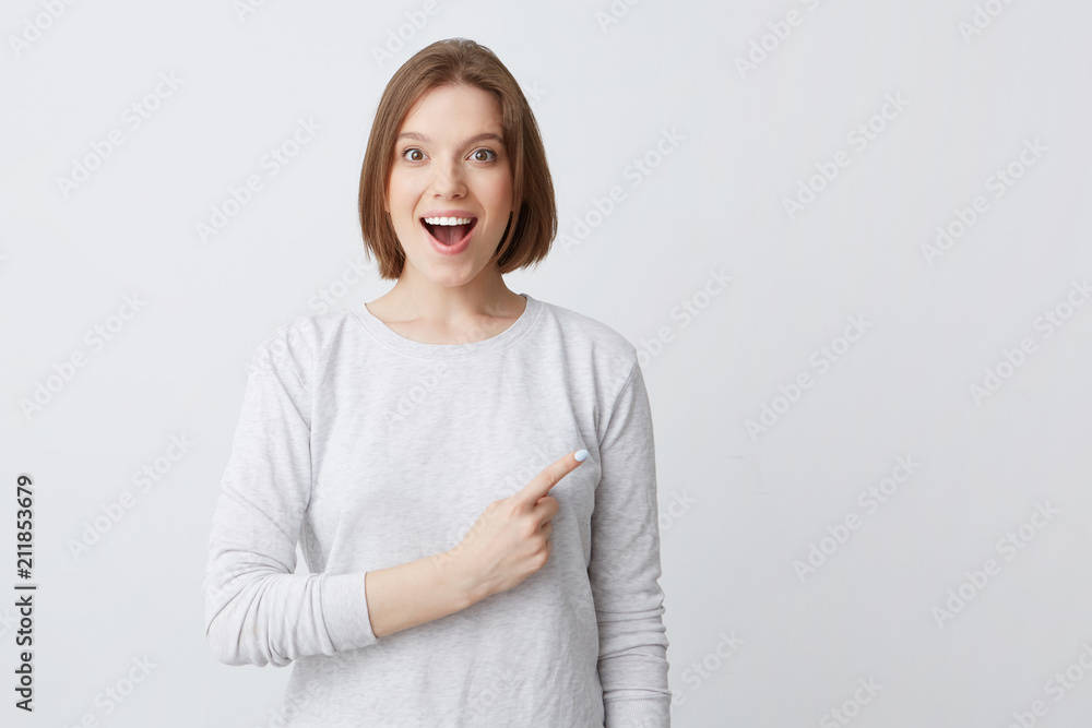 Fototapeta premium Surprised pretty young woman in longsleeve with opened mouth smiling and pointing to the side isolated over white background Feels happy and amazed