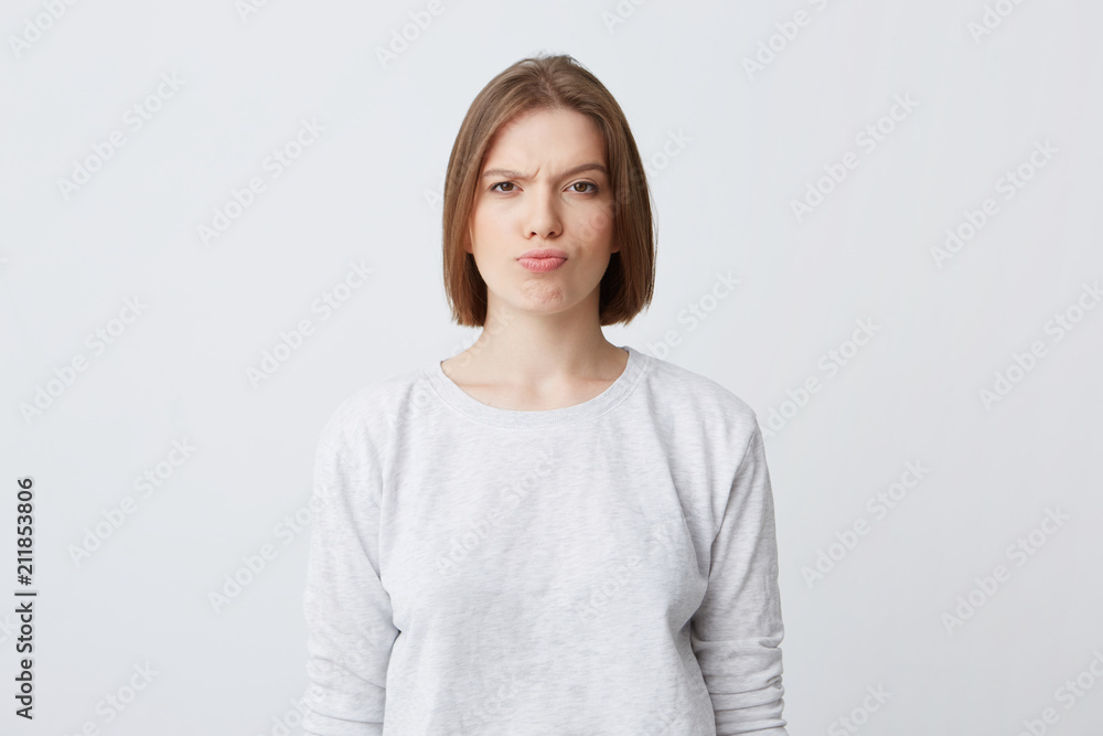 Portrait of serious unhappy young woman in longsleeve standing and frowning her face isolated over white background Looking directly in camera Portrait of serious unhappy young woman in longsleeve standing and frowning her face isolated over white background Looking directly in camera