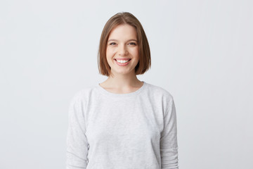 beautiful woman expresses happy emotions, has broad pleasant smile, wears white longsleeve and feels satisfied. Isolated over white background