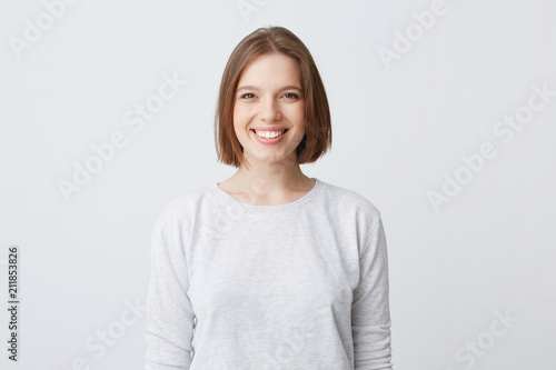 beautiful woman expresses happy emotions, has broad pleasant smile, wears white longsleeve and feels satisfied. Isolated over white background