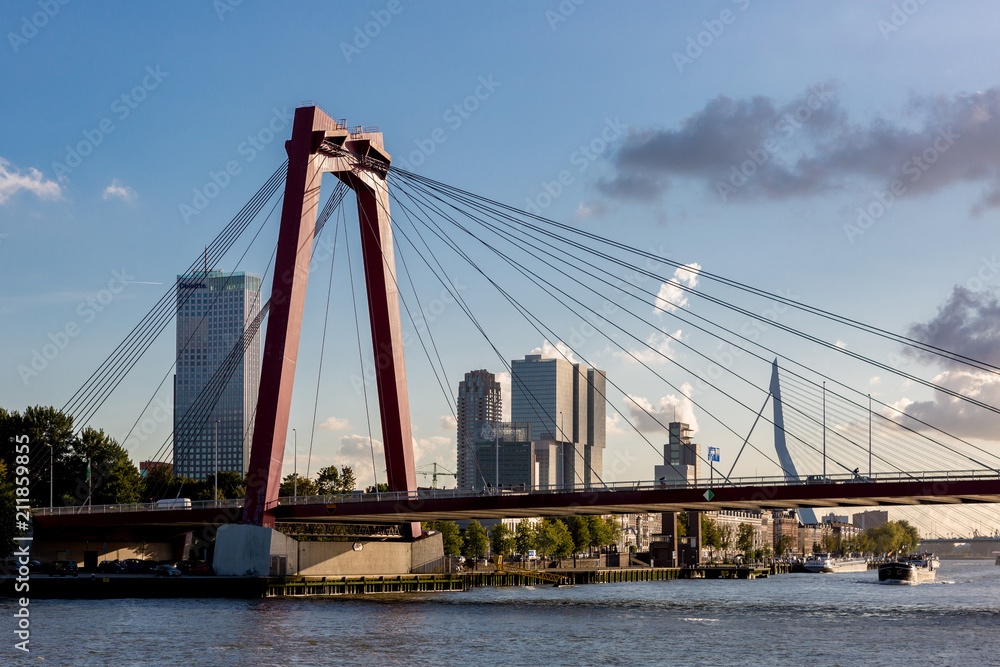 Orange cable bridge in Rotterdam with the cityscape of the Dutch city as a backdrop with the financial district and Erasmus bridge against a dramatic sunset sky with cumulus clouds over the skyline