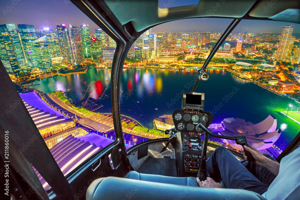 Fototapeta premium Wnętrze kokpitu helikoptera lecącego nad zatoką mariny w Singapurze z wieżowcami dzielnicy finansowej w nocy odbite w porcie. Lot widokowy nad panoramą Singapuru. Nocna scena miejska antena.