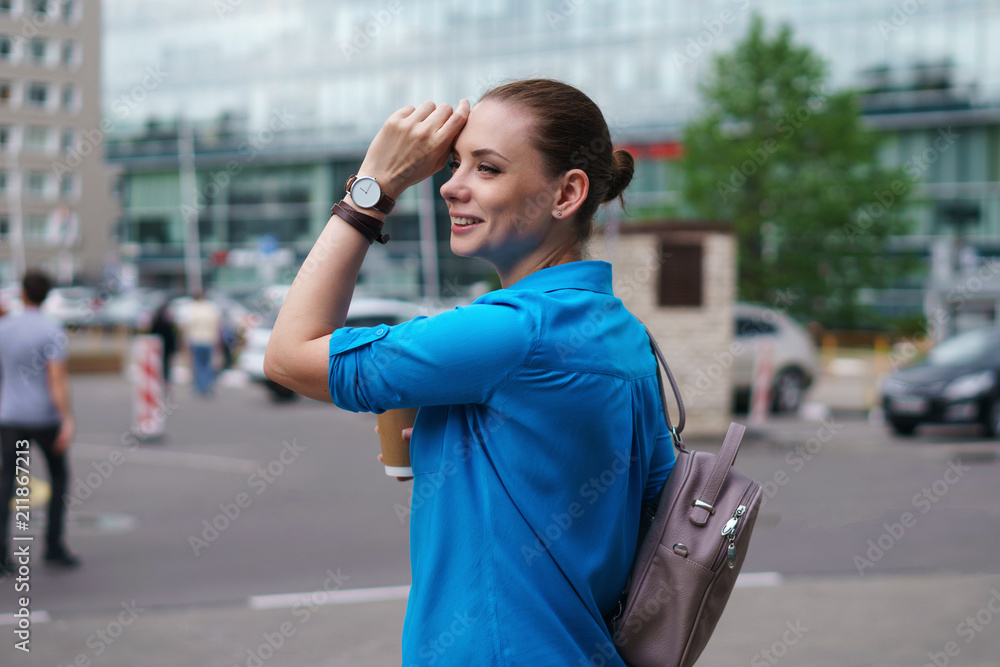 Young attractive girl with backpack holding cup of coffee. She stands on street of city. Woman straightens her hair and smiles. Young attractive girl with backpack holding cup of coffee. She stands on street of city. Woman straightens her hair and smiles.